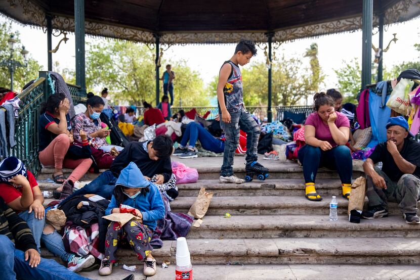 A boy plays with a roller-skate as other expelled migrants sit around a gazebo in a public...