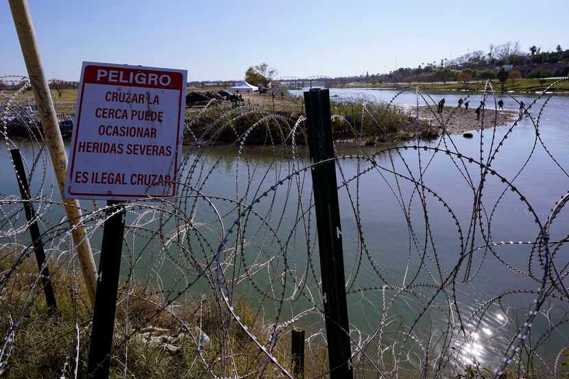 Migrants cross the Rio Grande into the U.S. from Mexico behind Concertina wire and a sign...