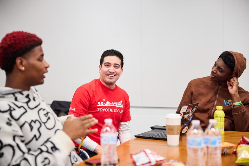 AN adult mentor in red shirt that says "Toyota Cares" sits at a table with two high school...