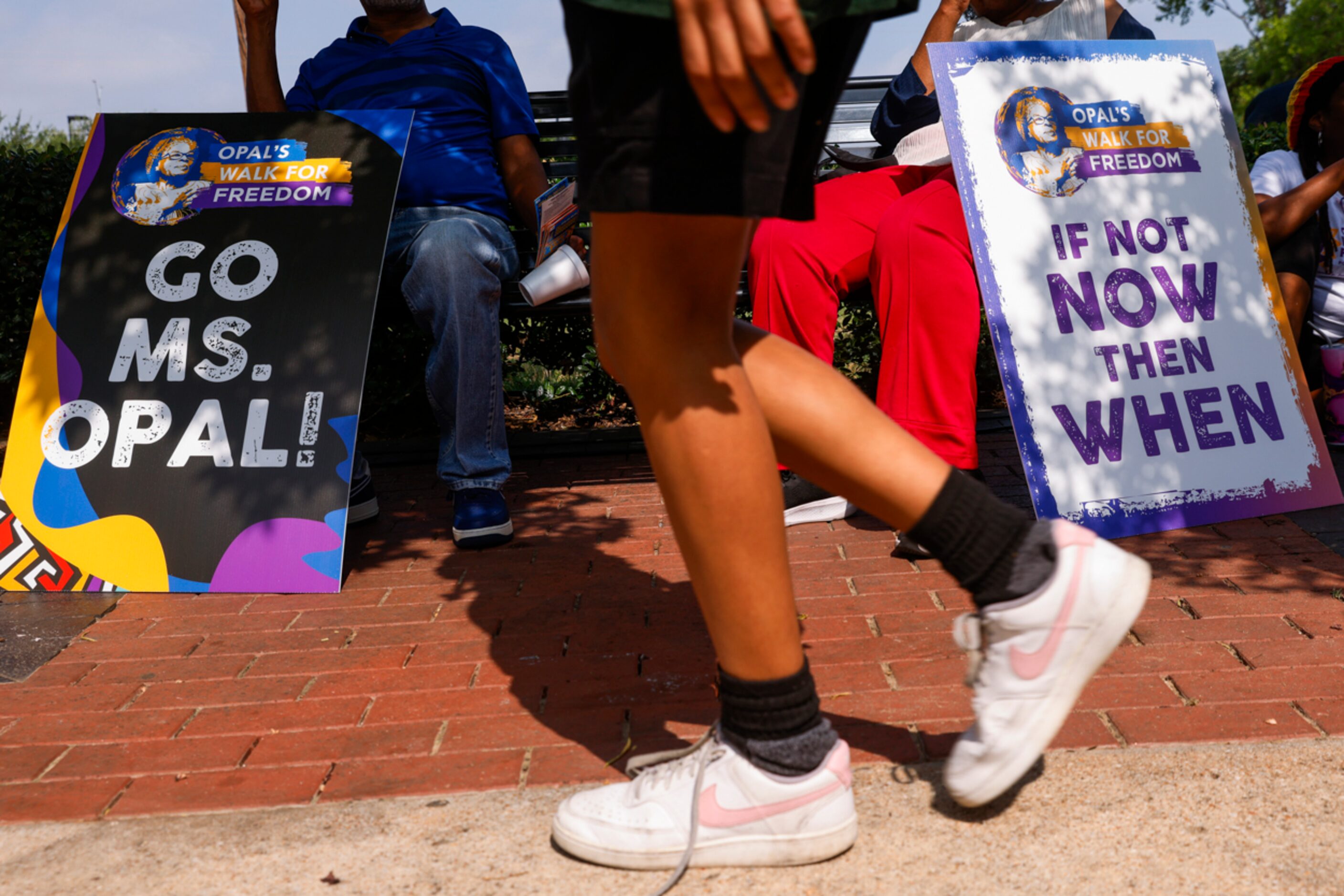 Attendees cool off under the shade, holding signs ahead of Opal's Walk for Freedom on...