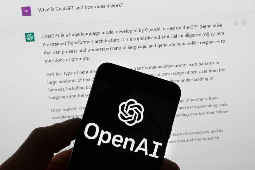 The OpenAI logo is seen on a mobile phone in front of a computer screen that displays output...