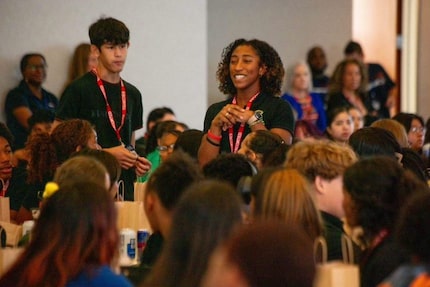 Two young people wearing lanyards stand in a crowded room surrounded by high school students...