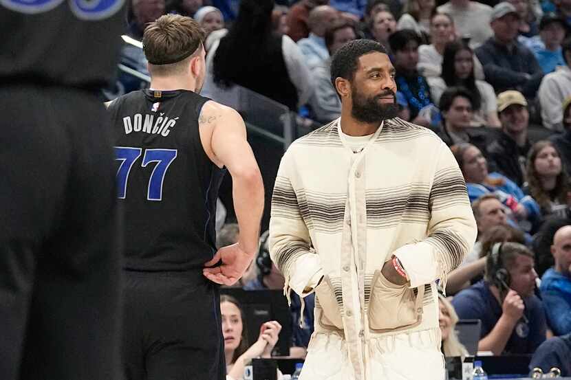 Injured Dallas Mavericks guard Kyrie Irving, right, looks on as teammate Luka Doncic (77)...