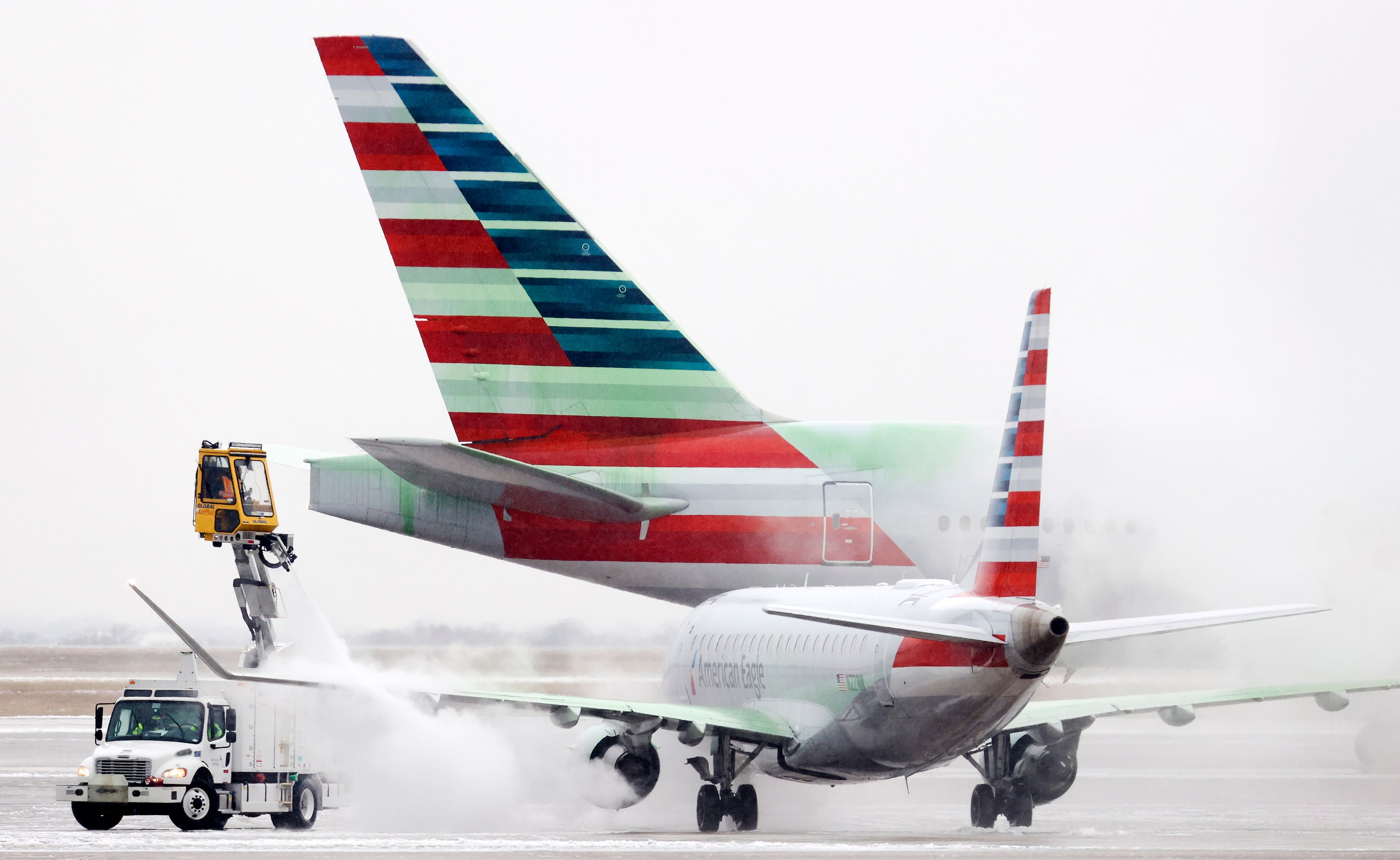 A deiced American Airlines Boeing 777 passes behind an American Embraer receiving the same...