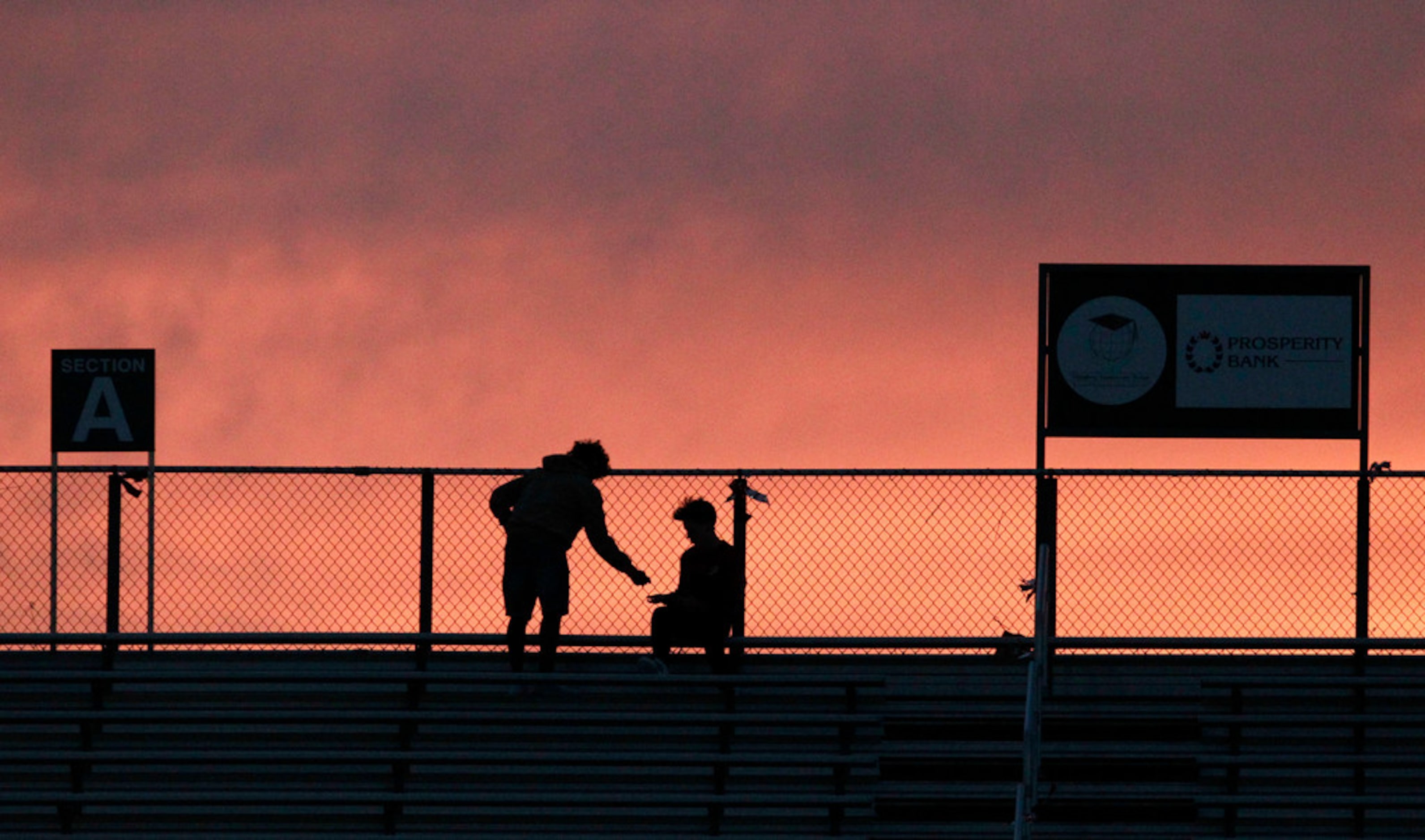 Two students talk high at the top of the stands in front of a pink sunset sky before the...