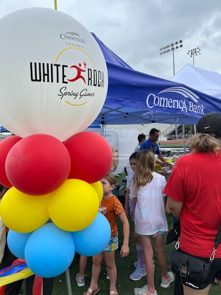 Kids stand next to balloons in front of a Comerica table at the White Rock Spring Games...