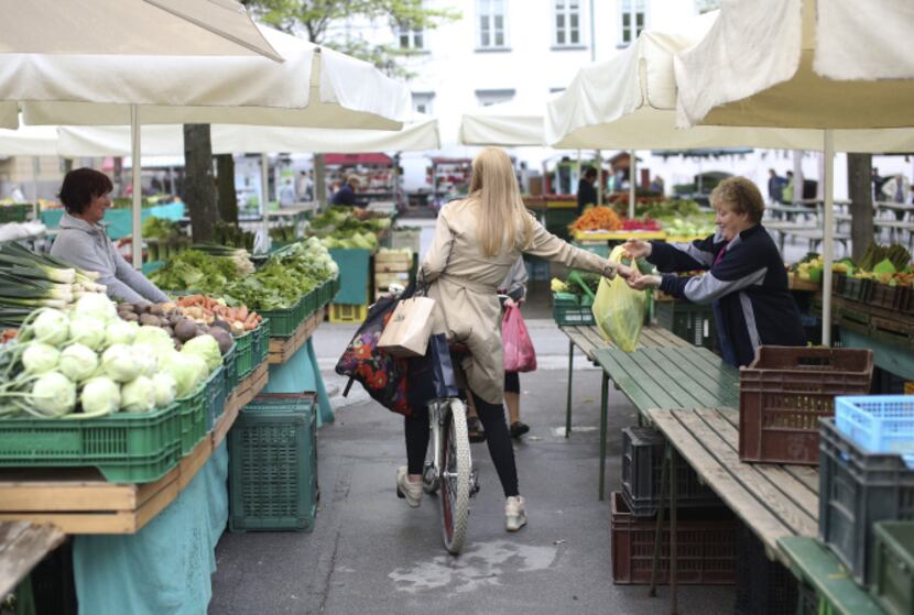 A customer pays a stallholder for a bag of produce while sitting on her bicycle at an open...