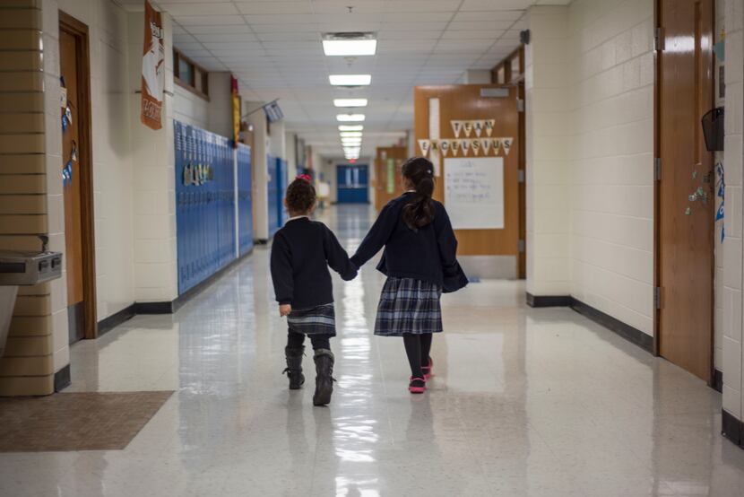 Two students in uniforms hold hands as they walk down the hallway of their school together.