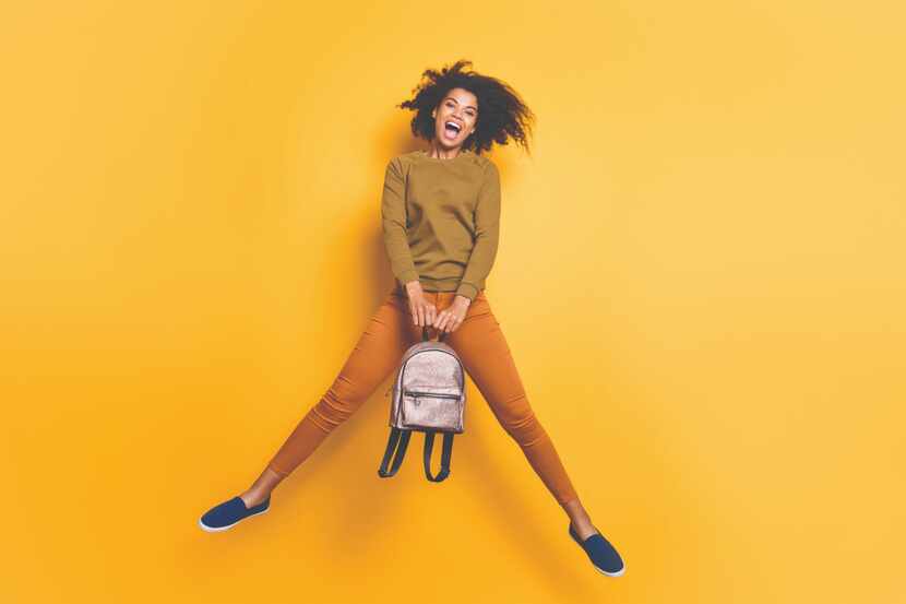 Full length photo of a cheerful, curly-haired student holding a handbag while jumping in the...