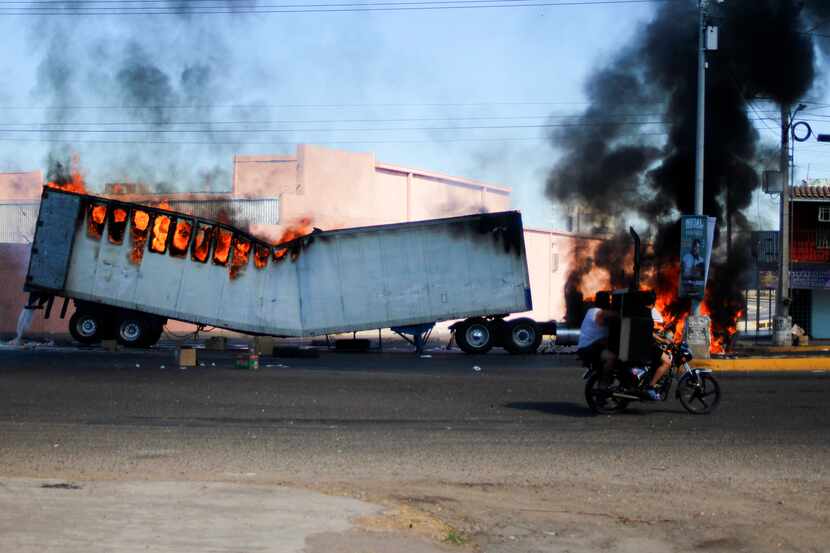 Dos hombres a bordo de una motocicleta pasan junto a un camión en llamas en las calles de...