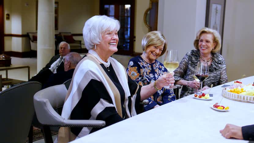 Three retiree women enjoy glasses of wine and snacks at a table together.