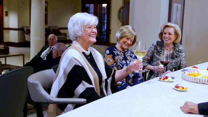 Three retiree women enjoy glasses of wine and snacks at a table together.