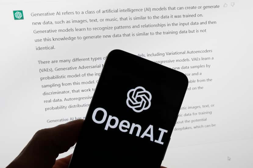 The OpenAI logo is seen on a mobile phone in front of a computer screen displaying output...