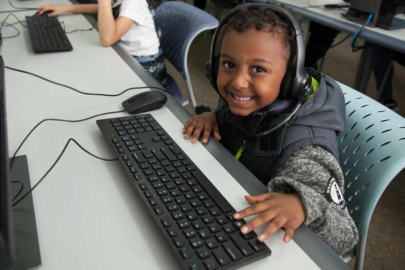 A child wearing a black vest and gray jacket uses a computer while wearing headphones and...