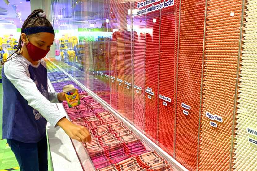 This photo shows a girl picking out crayons from a Crayon wall at a Crayola store.