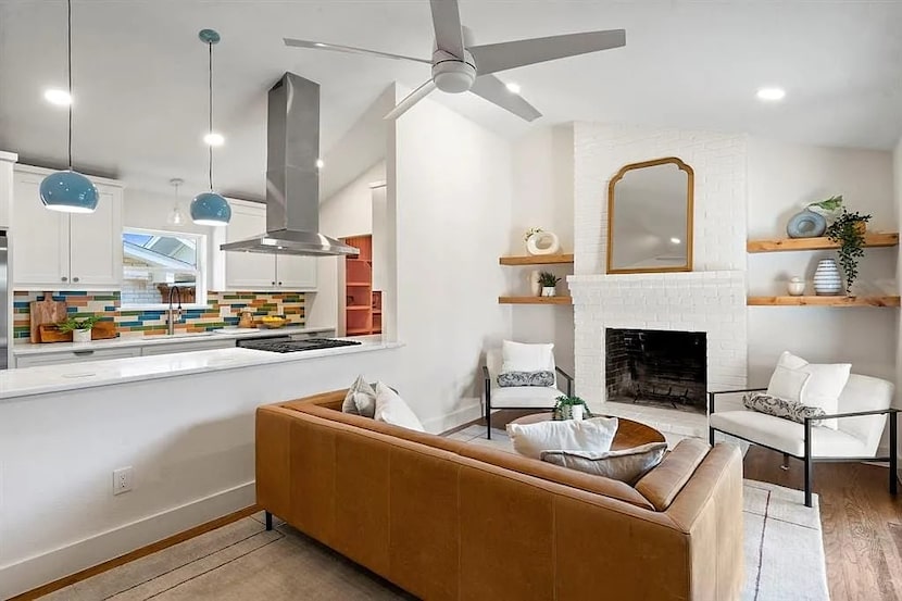 Living room with tan sofa and white chairs around a white brick fireplace, kitchen to the left