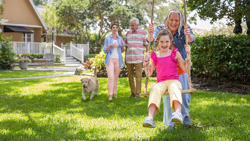 Grandmother pushes her laughing granddaughter on a swing outside while parents look on.