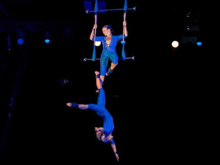 An acrobat in a show hangs on a swing suspended from the ceiling while another acrobat hangs...