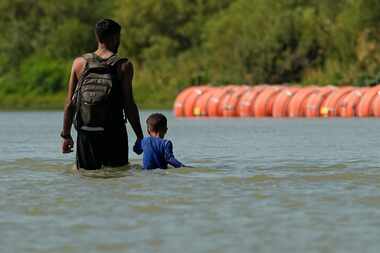 Migrants walk past large buoys being used as a floating border barrier on the Rio Grande,...