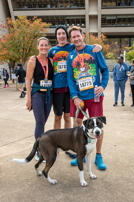 Three race attendees wearing race bibs pose with a dog on a leash.