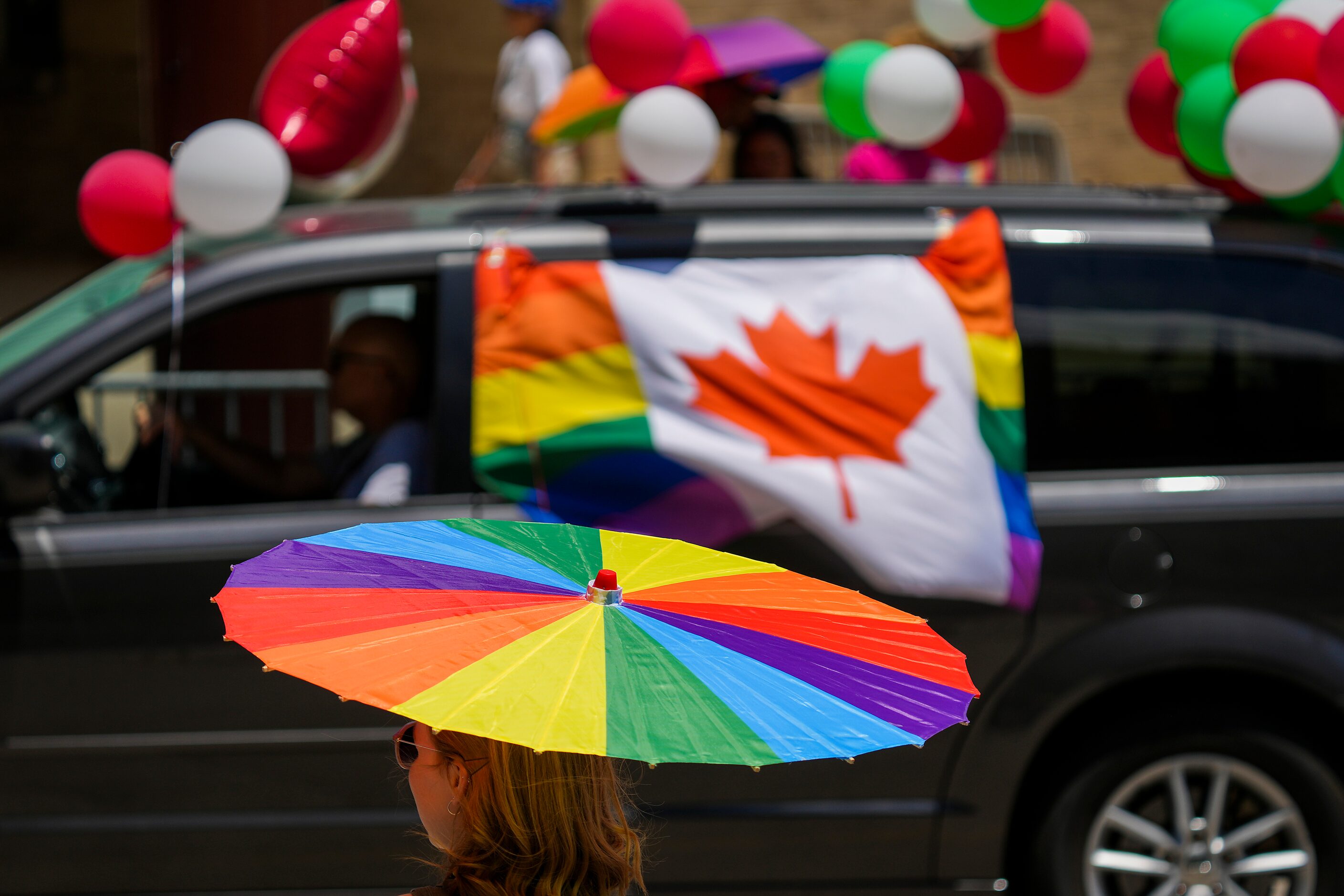 A spectator takes cover under a rainbow colored parasol as participants from Consulate...