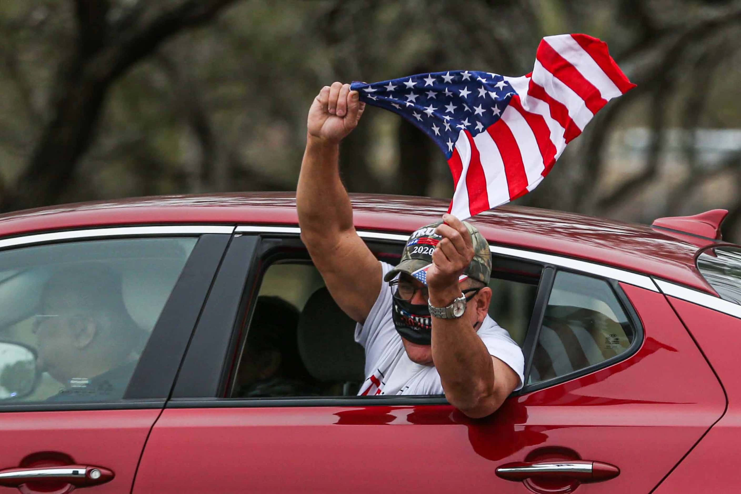 A man shouts from a car window in motion holding an American flag as he passes in front of a...