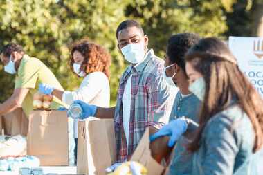 A man wearing a mask places a canned food item into a paper bag as he volunteers with his...