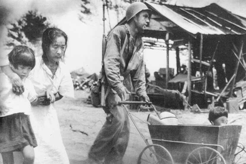 A battle-weary Marine leads a Saipan family toward a temporary home. From Their Backs...