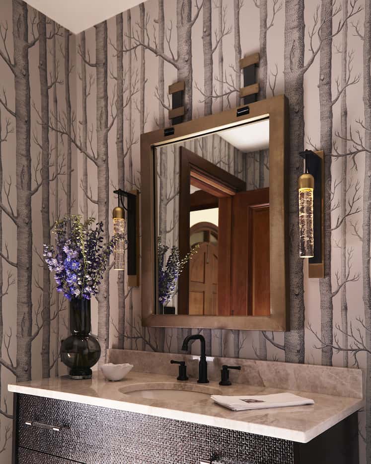Moody powder bath with tree-patterned wallpaper, a dark gold mirror flanked by sconces and a...
