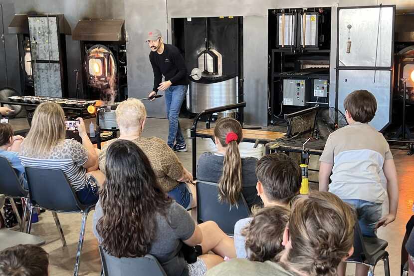 A man demonstrates glassblowing techniques to a crowd in a studio.