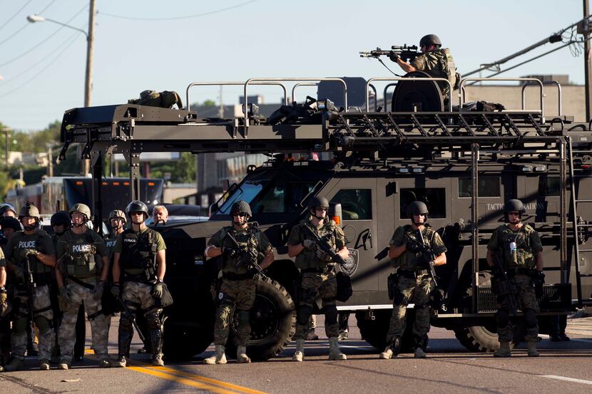 Law enforcement officers, including a sniper perched atop an armored vehicle, watch as...