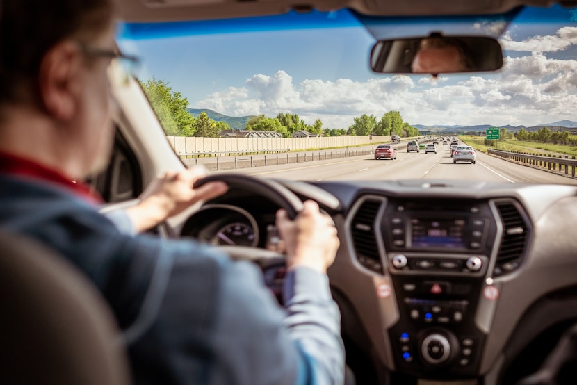 A man and the dashboard of his car defocused in the foreground, with light daytime traffic...