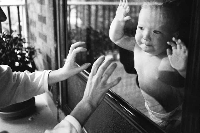 Separated by a window pane, a mother playfully taps on the glass while her child watches.
