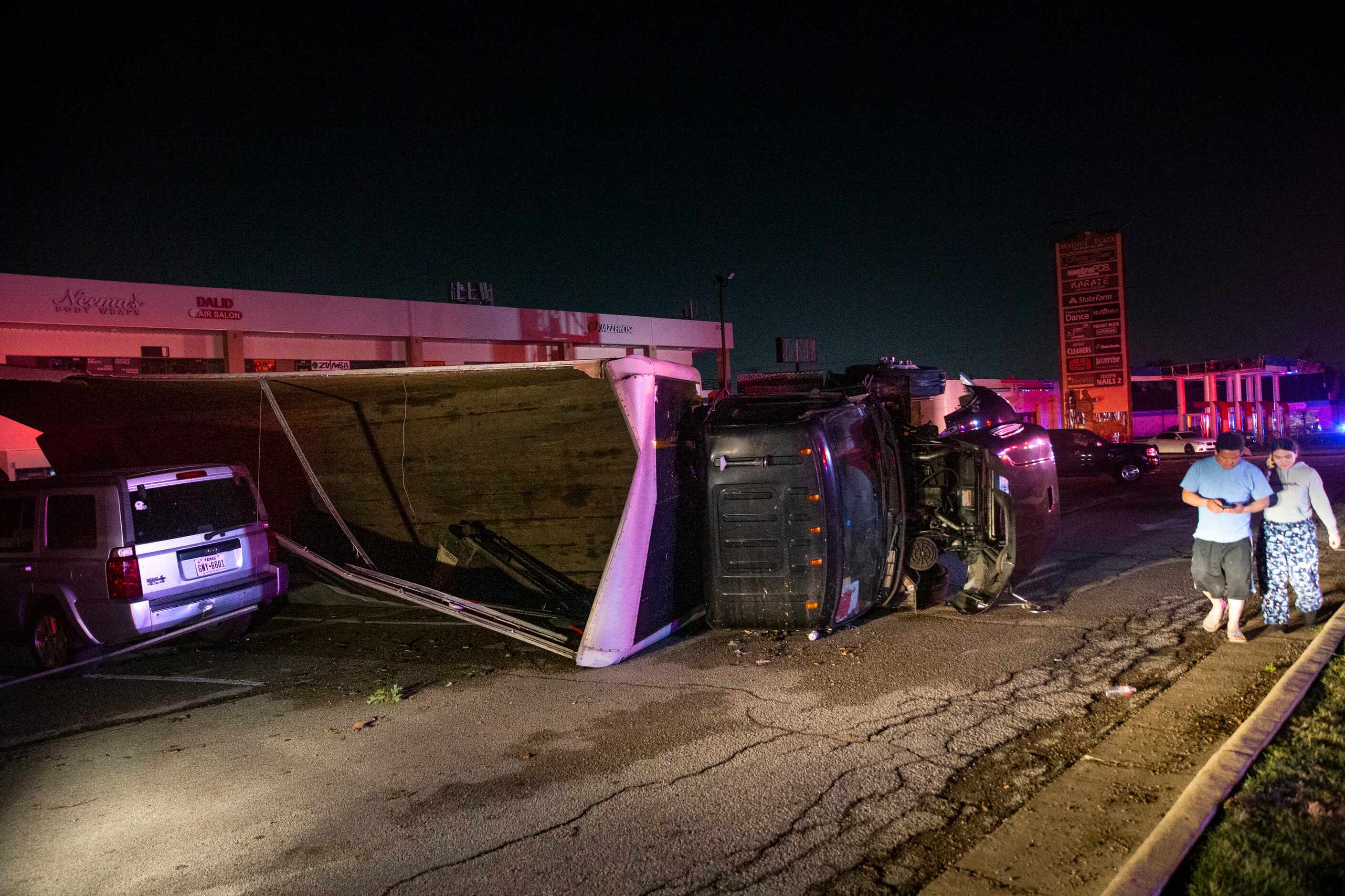 People walk past an overturned semi-truck at a shopping center on the corner of Walnut Hill...