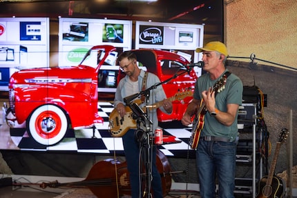 Two men play guitars and sing into microphones on a makeshift stage inside a garage space.