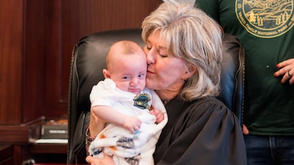 A female judge with gray hair sits at her judicial bench and kisses an infant child she is...