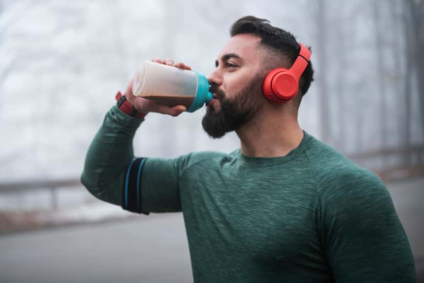 A man drinks a protein shake from a bottle while wearing a green sweater post-workout