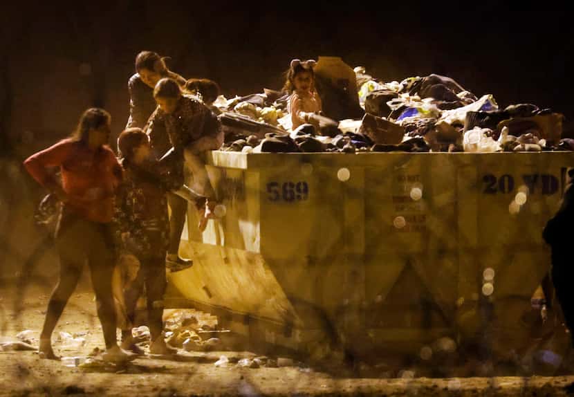 Young migrant children rummage through large trash dumpsters of discarded blankets,...