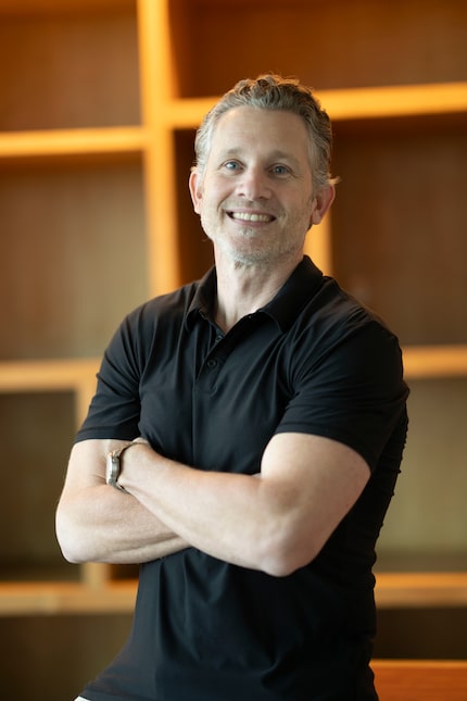 A man in a black t-shirt with his arms crossed over his chest smiles at the camera.