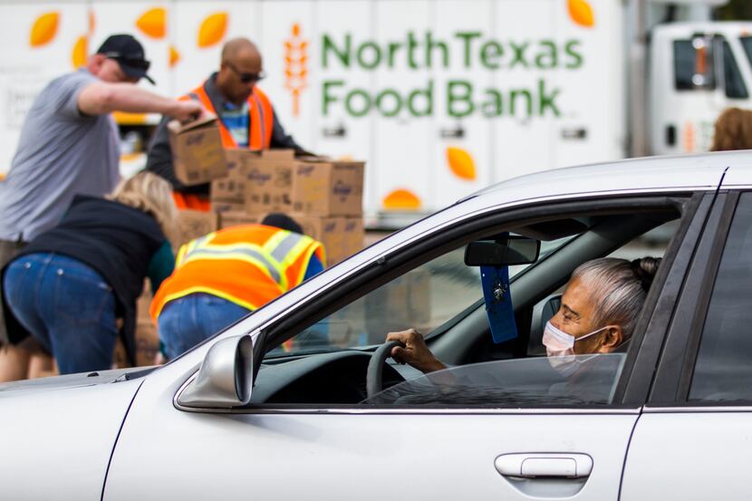 A woman wearing a surgical mask drives past volunteers who prepare to hand out boxes of food...