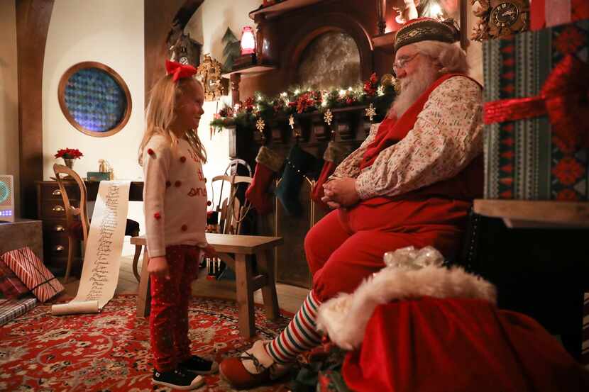 Foto de una niña rubia hablando con Santa en un cuarto que parece la casa de este, con...