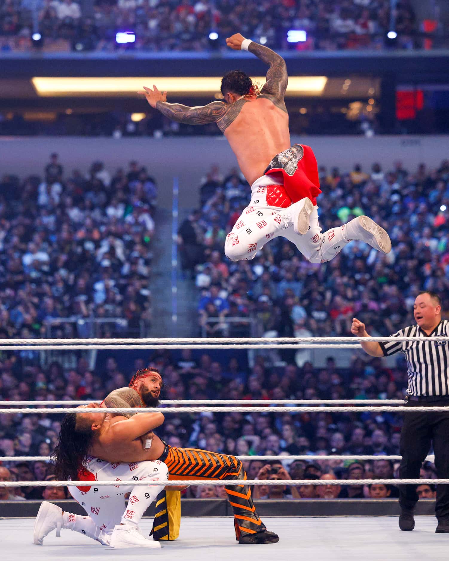Jey Uso leaps from the top rope while Jimmy Uso (white pants) holds down Shinsuke Nakamura...