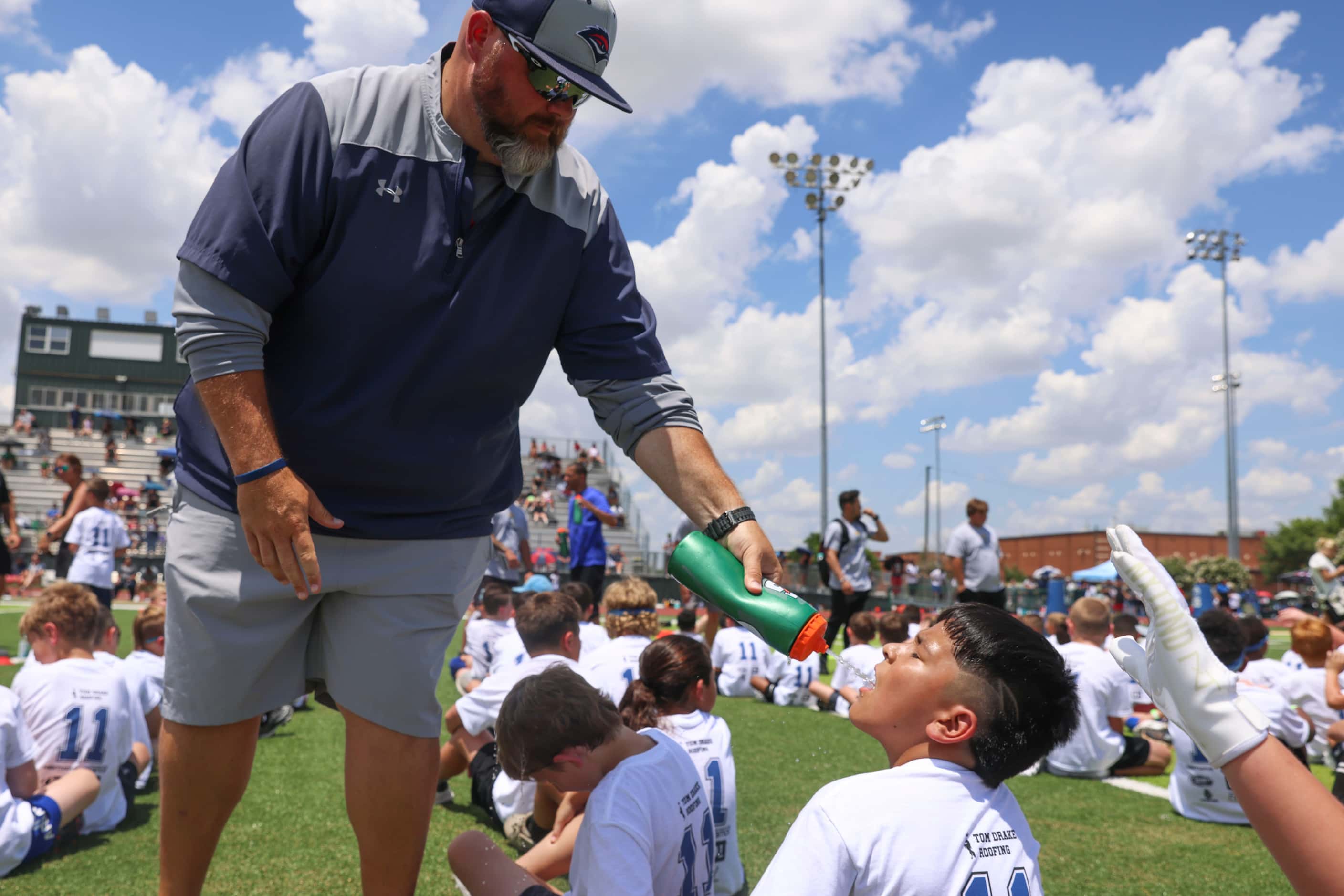 Coach Nick Brown sprays water on young football players as they line up to practice and...