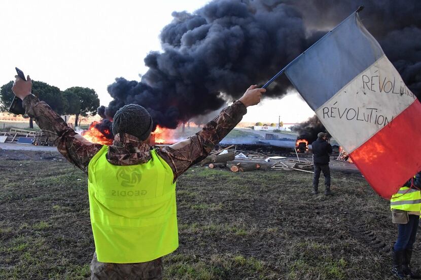 A man wearing a yellow vest waves a French flag in front of burning tires at a road blockade...