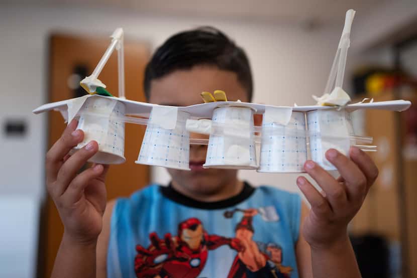 A young boy shows off a bridge he created using a variety of items such as cups, paper and...