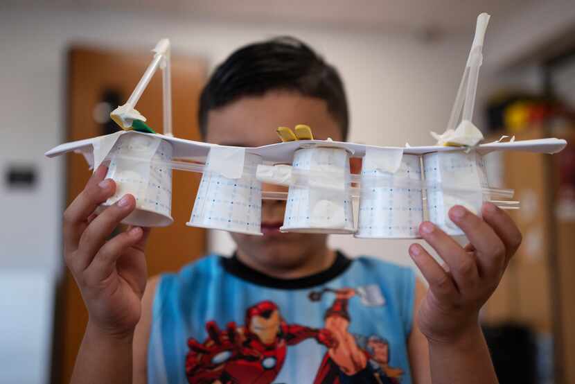 A young boy shows off a bridge he created using a variety of items such as cups, paper and...