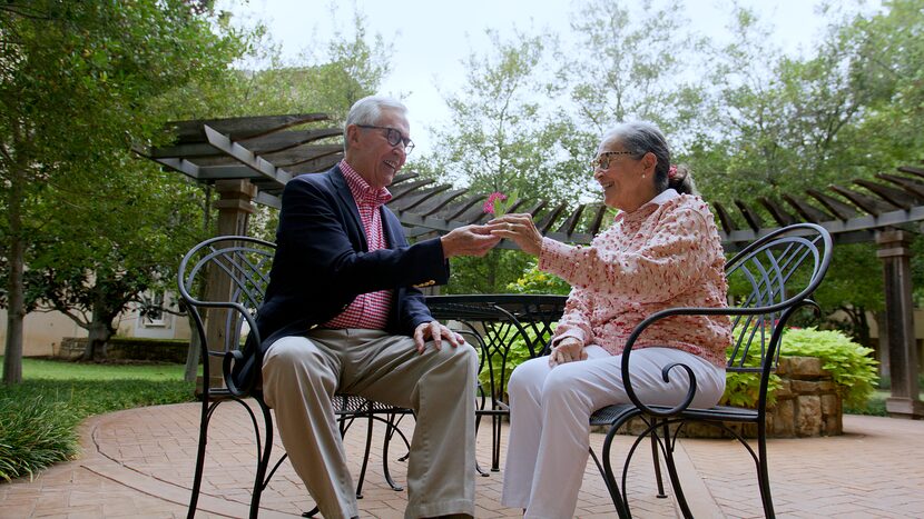 A man and women sit together at a table at an outdoor patio table, smiling as they pose with...