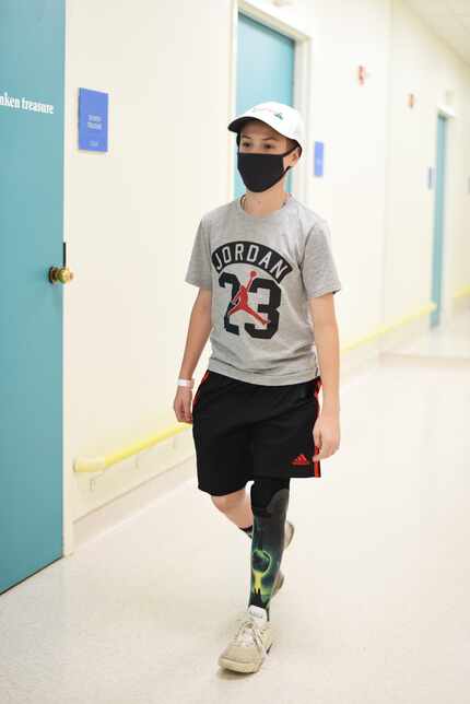 12-year-old Austin walks with his prothesis while wearing a mask and a gray shirt that reads...