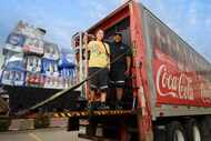 Two people stand on the loading apparatus of a Coca-Cola shipping truck.