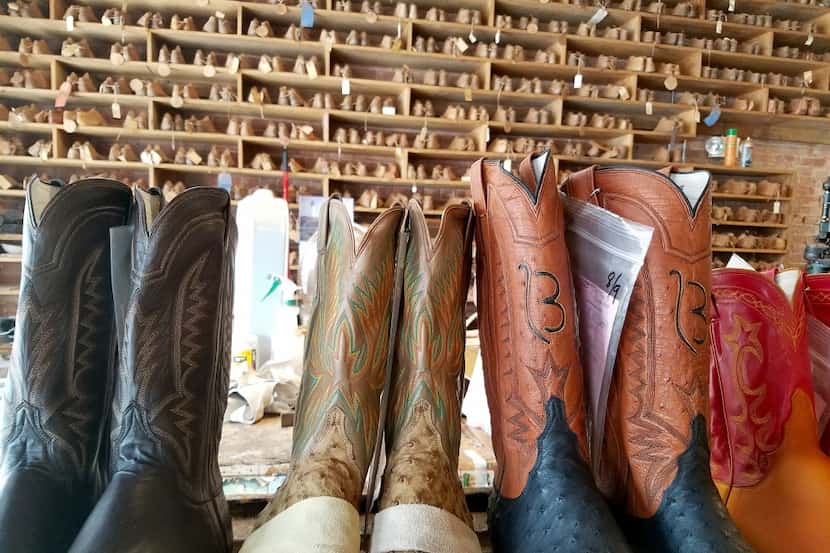 Boots in progress stand in front of a wall full of different sizes of boot foot forms at...
